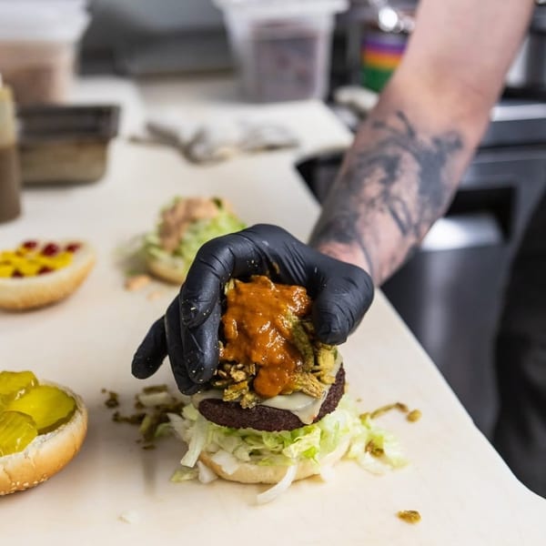 A gloved hand adds sauce and crispy toppings to a burger with lettuce and cheese.
