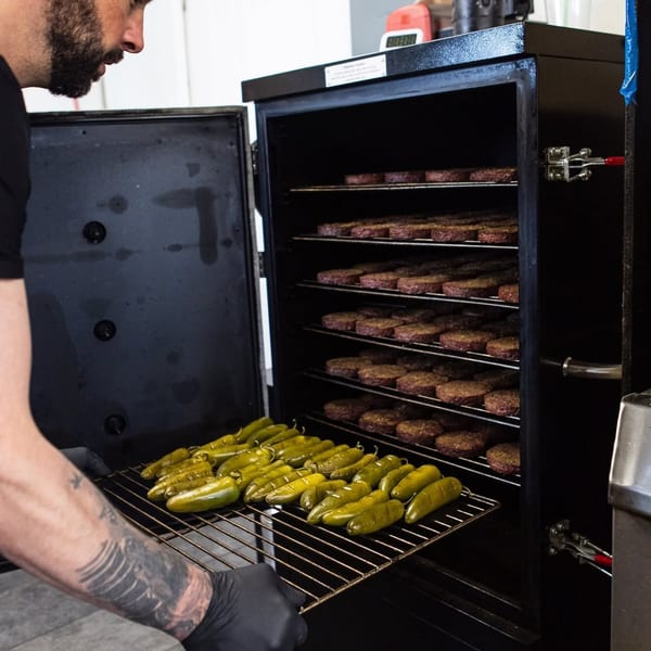 Chef placing a tray of green peppers into a smoker filled with burger patties.