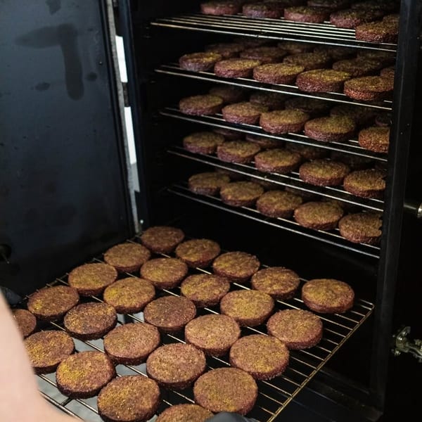 A chef in black gloves places a rack of seasoned burger patties into a large smoker.