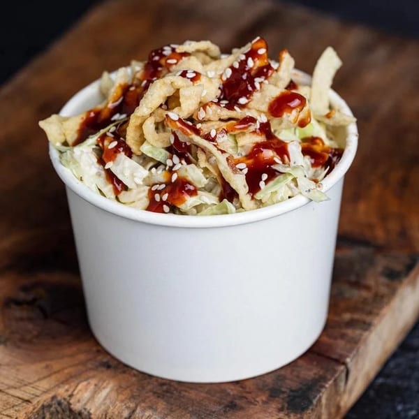 Asian slaw with crispy wontons, sesame seeds, and sweet chili sauce in a white cup.