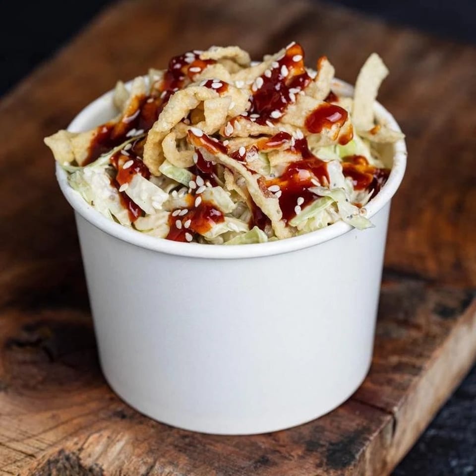 Asian slaw with crispy wontons, sesame seeds, and sweet chili sauce in a white cup.