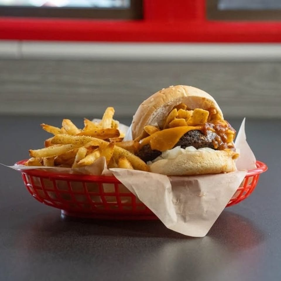 A chili cheeseburger with Fritos and onions, served with fries in a red basket.