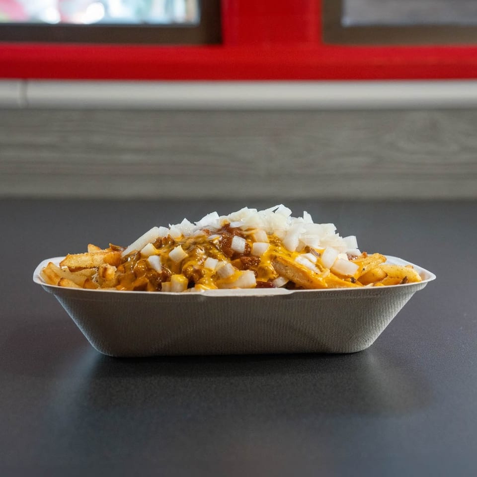 Chili cheese fries topped with diced white onions in a white paper tray on a dark surface.