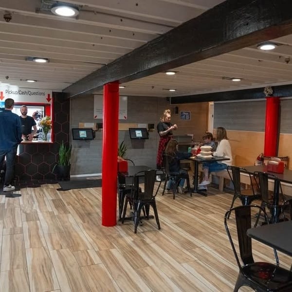 Restaurant interior with customers at tables and a pickup counter, featuring red accents and wood-look flooring.