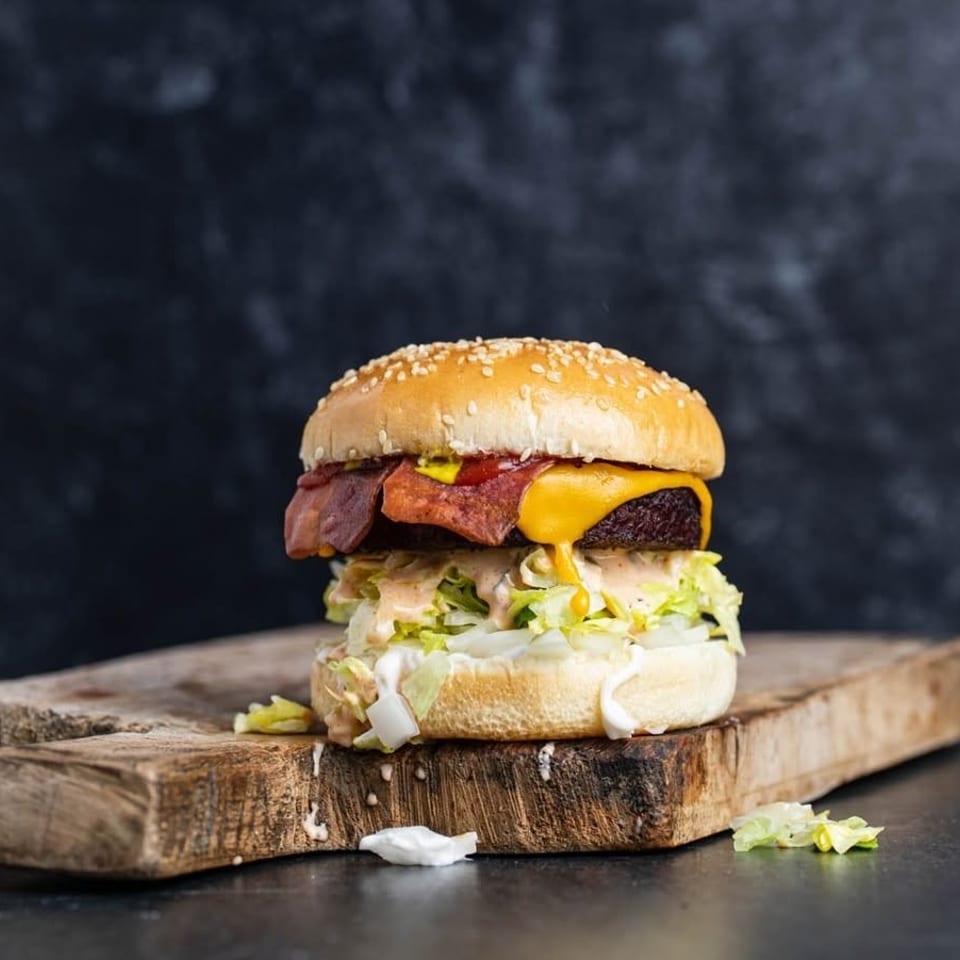 A juicy burger with melted cheese, bacon, lettuce, and sauce on a wooden board.